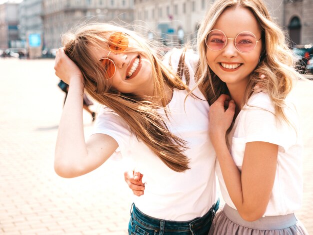 Un ritratto di due giovani belle ragazze sorridenti bionde dei pantaloni a vita bassa in vestiti bianchi della maglietta dell'estate d'avanguardia.