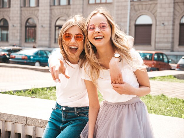 Un ritratto di due giovani belle ragazze sorridenti bionde dei pantaloni a vita bassa in vestiti bianchi della maglietta dell'estate d'avanguardia.