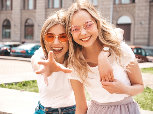 Un ritratto di due giovani belle ragazze sorridenti bionde dei pantaloni a vita bassa in vestiti bianchi della maglietta dell'estate d'avanguardia.