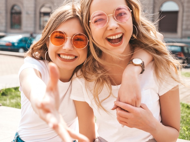 Un ritratto di due giovani belle ragazze sorridenti bionde dei pantaloni a vita bassa in vestiti bianchi della maglietta dell'estate d'avanguardia.