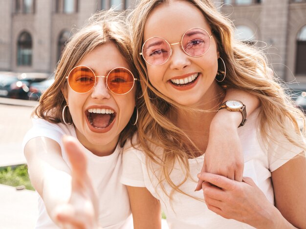 Un ritratto di due giovani belle ragazze sorridenti bionde dei pantaloni a vita bassa in vestiti bianchi della maglietta dell'estate d'avanguardia.