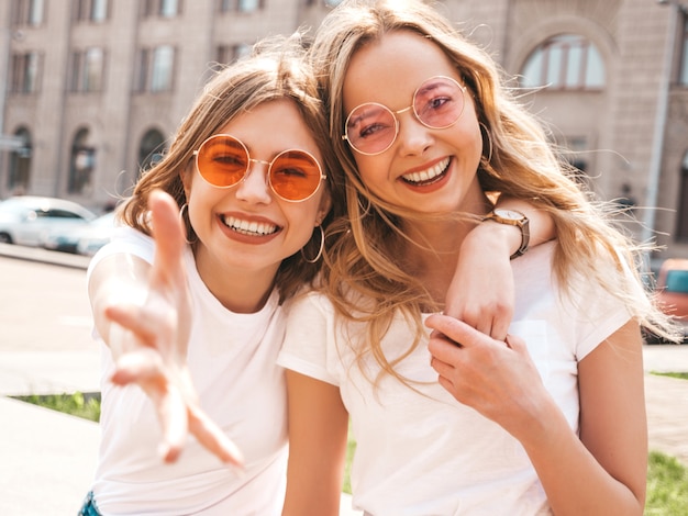 Un ritratto di due giovani belle ragazze sorridenti bionde dei pantaloni a vita bassa in vestiti bianchi della maglietta dell'estate d'avanguardia.