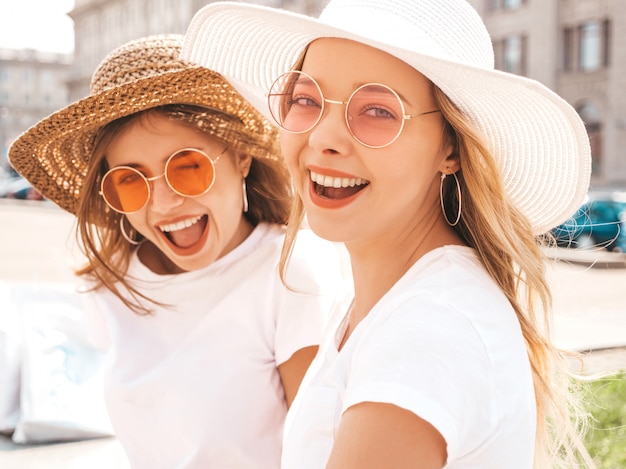 Un ritratto di due giovani belle ragazze sorridenti bionde dei pantaloni a vita bassa in vestiti bianchi della maglietta dell'estate d'avanguardia.