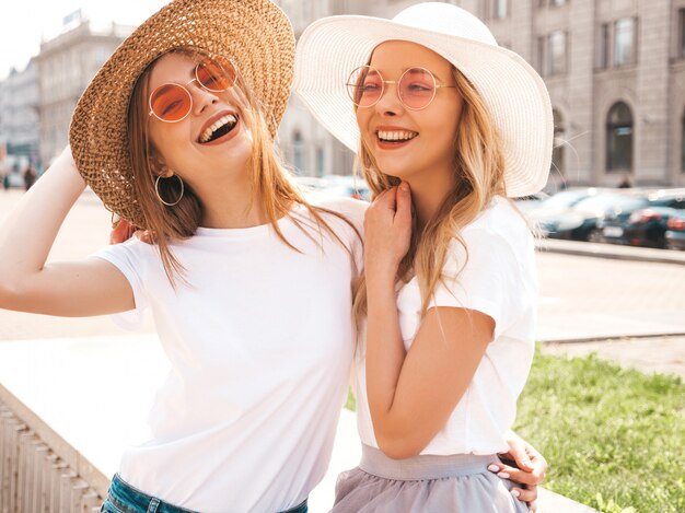 Un ritratto di due giovani belle ragazze sorridenti bionde dei pantaloni a vita bassa in vestiti bianchi della maglietta dell'estate d'avanguardia.