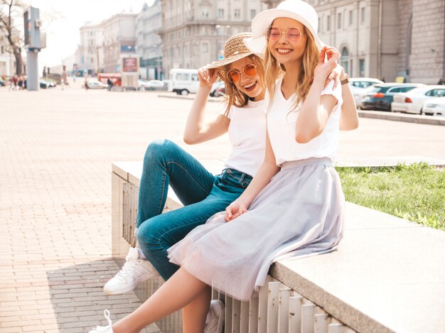 Un ritratto di due giovani belle ragazze sorridenti bionde dei pantaloni a vita bassa in vestiti bianchi della maglietta dell'estate d'avanguardia.