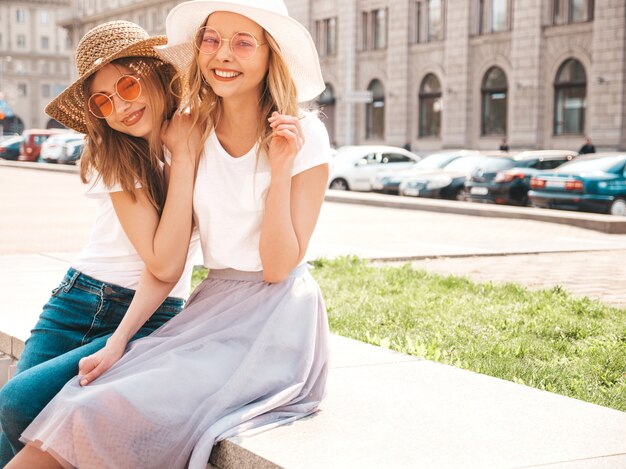 Un ritratto di due giovani belle ragazze sorridenti bionde dei pantaloni a vita bassa in vestiti bianchi della maglietta dell'estate d'avanguardia.