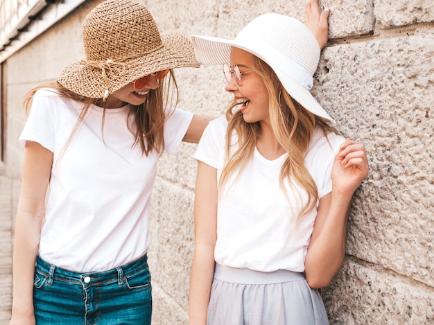 Un ritratto di due giovani belle ragazze sorridenti bionde dei pantaloni a vita bassa in vestiti bianchi della maglietta dell'estate d'avanguardia.