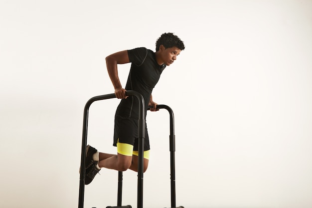 Un ritratto di un giovane afroamericano muscolare concentrato in abiti da allenamento neri facendo tuffi sulle barre parallele su bianco