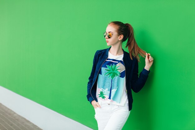 Un ritratto di una giovane ragazza all'aperto vicino al muro verde con una linea bianca verso il basso. La ragazza indossa occhiali da sole, tiene in mano la coda dei capelli e guarda lontano.