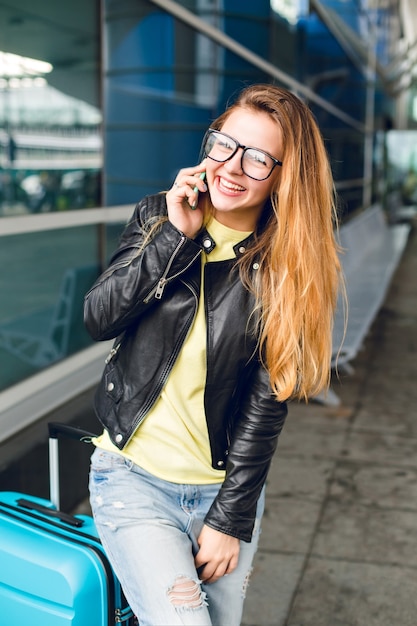 Un ritratto orizzontale di una bella ragazza con i capelli lunghi in piedi fuori in aeroporto. Indossa maglione giallo, giacca nera e jeans. Sta parlando al telefono e sorride alla telecamera.