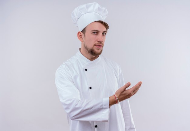 Un serio giovane barbuto chef uomo che indossa l'uniforme da cucina bianca e un cappello che invita a venire con la mano mentre guarda un muro bianco