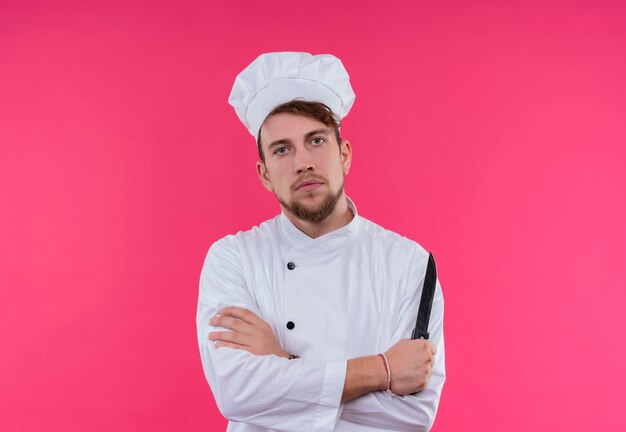 Un serio giovane barbuto uomo chef in uniforme bianca che tiene un coltello mentre guarda su una parete rosa