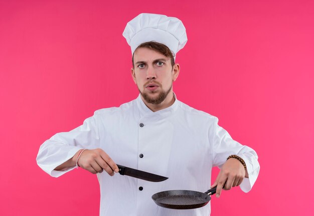 Un serio giovane barbuto uomo chef in uniforme bianca tenendo la padella con il coltello mentre guarda su una parete rosa