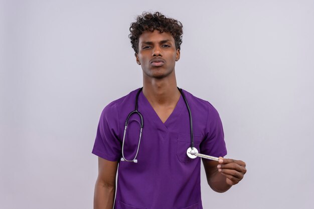 Un serio giovane medico dalla carnagione scura bello con capelli ricci che indossa l'uniforme viola con lo stetoscopio mentre tiene il termometro