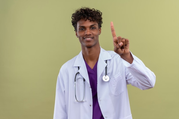Un sorridente giovane bello di carnagione scura medico con i capelli ricci che indossa camice bianco con uno stetoscopio che mostra il numero uno con il dito indice su uno spazio verde
