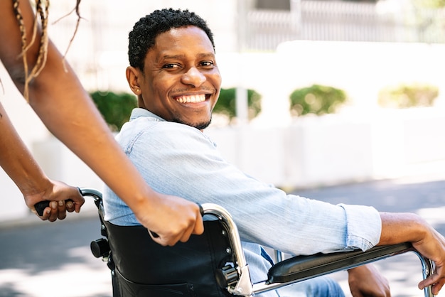 Un uomo afroamericano su una sedia a rotelle sorride e guarda la telecamera mentre la sua ragazza lo spinge.