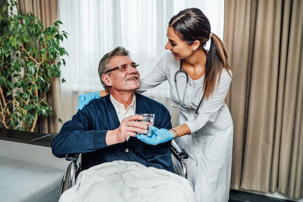 Un uomo anziano su una sedia a rotelle sorride all'infermiera, lei gli porge un bicchiere d'acqua.