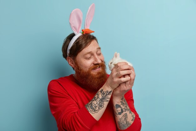 Un uomo barbuto e allegro e premuroso gioca con un coniglietto carino, indossa orecchie di coniglio e maglione rosso, celebra la Pasqua, si gode la primavera, posa al coperto. Tradizioni e concetto di feste religiose