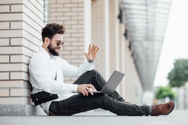 Un uomo barbuto, felice, sorridente ed elegante seduto sulla farina per le strade della città vicino al moderno centro uffici e che lavora con il suo laptop