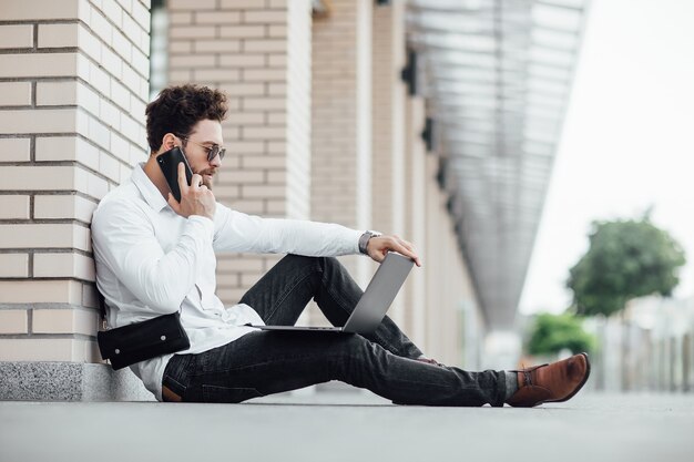 Un uomo barbuto, felice, sorridente ed elegante seduto sulla farina per le strade della città vicino al moderno centro uffici e lavora con il suo laptop e chiama il telefono