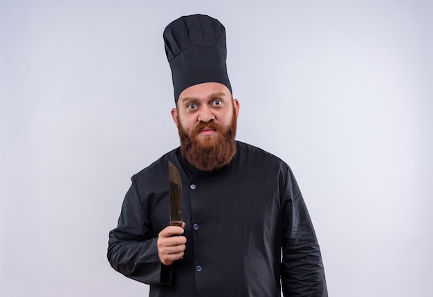 Un uomo barbuto serio chef in uniforme nera che tiene il coltello mentre guarda la fotocamera su un muro bianco