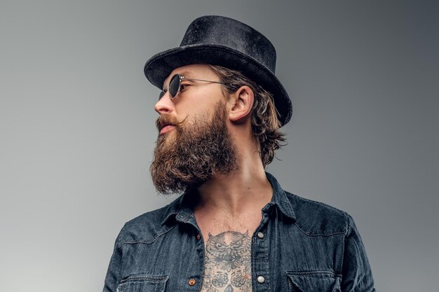 Un uomo barbuto serio con cappello e occhiali da sole sta posando in studio fotografico.