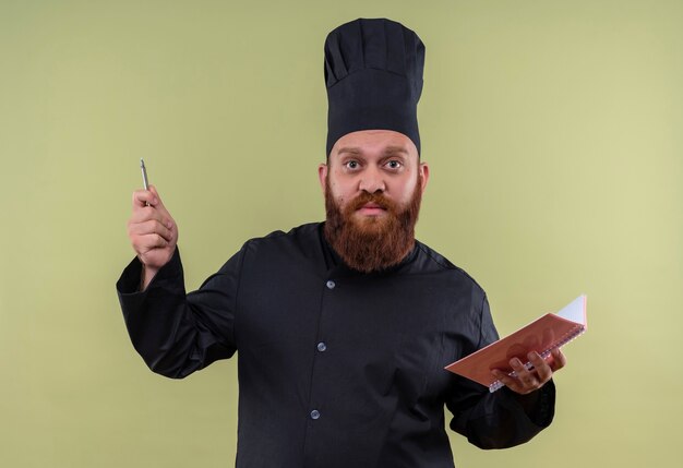 Un uomo barbuto serio del cuoco unico in taccuino e matita della tenuta dell'uniforme nera mentre osserva e prova a spiegare qualcosa su una parete verde