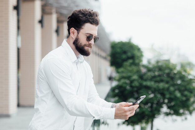 Un uomo barbuto, serio ed elegante in camicia bianca e occhiali da sole in piedi per le strade della città e navigando su smartphone