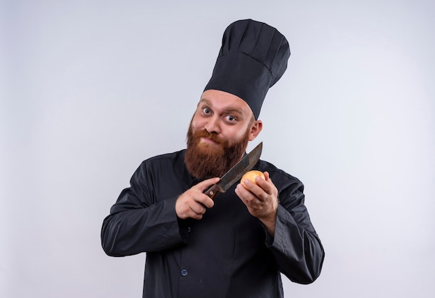 Un uomo chef barbuto felice in uniforme nera che cerca di tagliare la cipolla con un coltello mentre guarda la fotocamera su un muro bianco