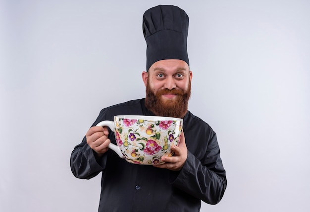 Un uomo chef barbuto felice in uniforme nera che tiene una tazza enorme di grandi dimensioni mentre guarda la fotocamera su un muro bianco