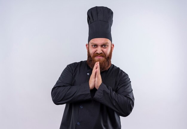 Un uomo chef barbuto stressante in uniforme nera che tiene le mani insieme mentre guarda su un muro bianco