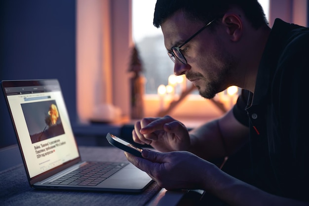 Un uomo con uno smartphone si siede davanti a un laptop a tarda notte
