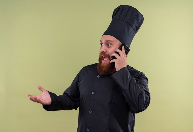 Un uomo di chef barbuto sorpreso in uniforme nera, parlando al telefono cellulare su una parete verde