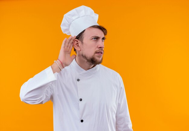 Un uomo dispiaciuto giovane chef barbuto in uniforme bianca che cerca di ascoltare con la mano sull'orecchio su una parete arancione