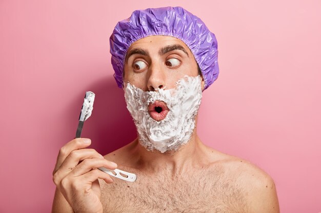 Un uomo europeo scioccato tiene la lama da barba, applica gel schiumogeno sulle guance, si rade le setole, indossa un cappuccio protettivo viola, ha una routine igienica