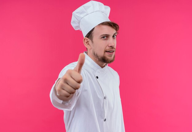 Un uomo felice giovane chef barbuto in uniforme bianca che mostra entrambi i pollici in su mentre guarda su una parete rosa