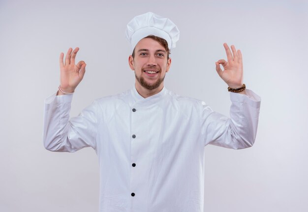 Un uomo felice giovane chef barbuto in uniforme bianca che mostra il gesto giusto con due mani su una parete bianca