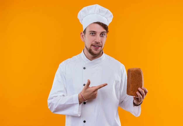 Un uomo felice giovane chef barbuto in uniforme bianca che punta a una pagnotta di pane con il dito indice su una parete arancione