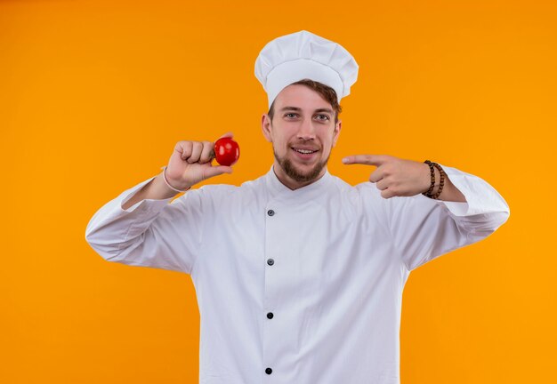 Un uomo felice giovane chef barbuto in uniforme bianca che punta al pomodoro fresco con il dito indice su una parete arancione