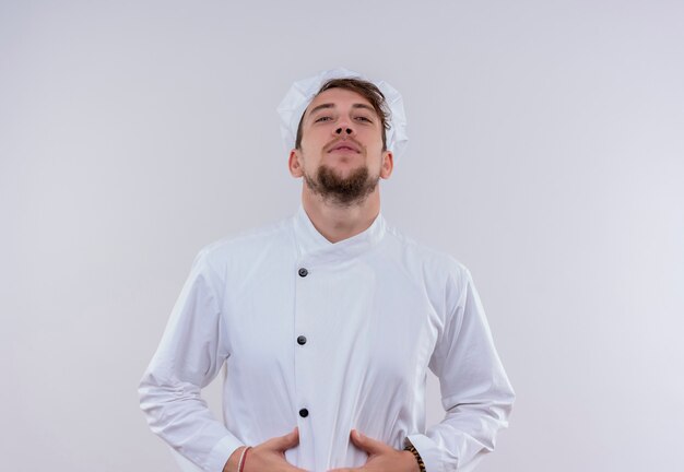 Un uomo giovane chef barbuto in uniforme bianca che tiene la mano sulla pancia mentre guarda su un muro bianco