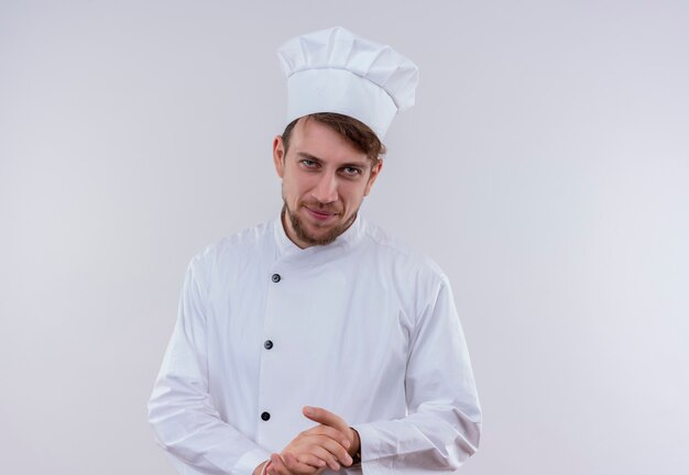 Un uomo giovane chef barbuto in uniforme bianca che tiene le mani insieme mentre guarda su un muro bianco