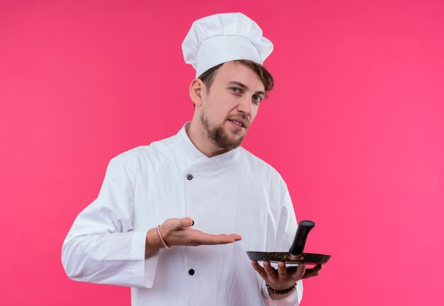 Un uomo giovane chef barbuto scontento in uniforme bianca che mostra la padella mentre guarda su una parete rosa