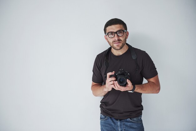 Un uomo giovane hipster in oculari detiene una fotocamera DSLR nelle mani in piedi contro un muro bianco