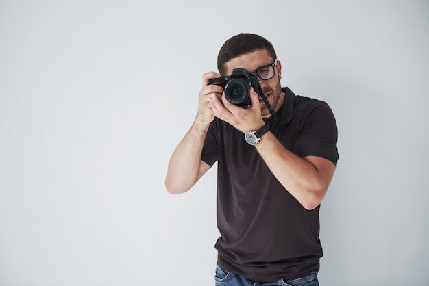 Un uomo giovane hipster in oculari detiene una fotocamera DSLR nelle mani in piedi contro un muro bianco