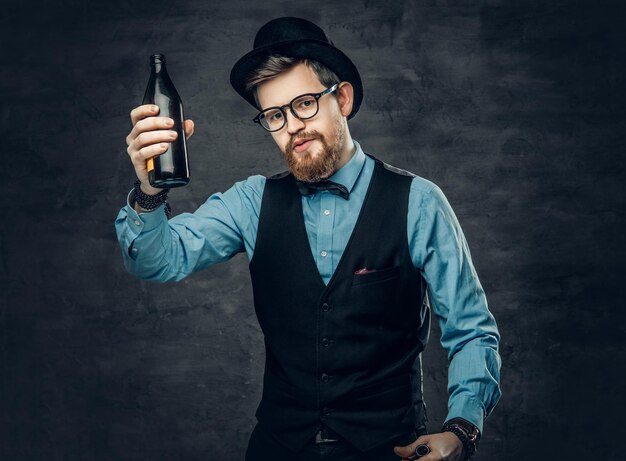 Un uomo hipster barbuto funky vestito con una camicia blu, un gilet elegante e un cappello a cilindro tiene una bottiglia di birra artigianale.