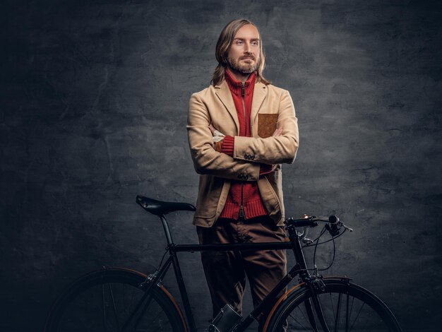 Un uomo hipster viaggiatore con lunghi capelli biondi, che indossa una giacca gialla in posa con un'autentica bicicletta a velocità singola.