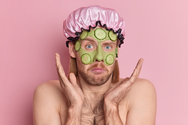 Un uomo incapace con barba ispida e baffi alza i palmi sul viso si sente esitante applica una maschera di bellezza verde con fette di cetriolo in piedi spalle nude indossa cappello da bagno.