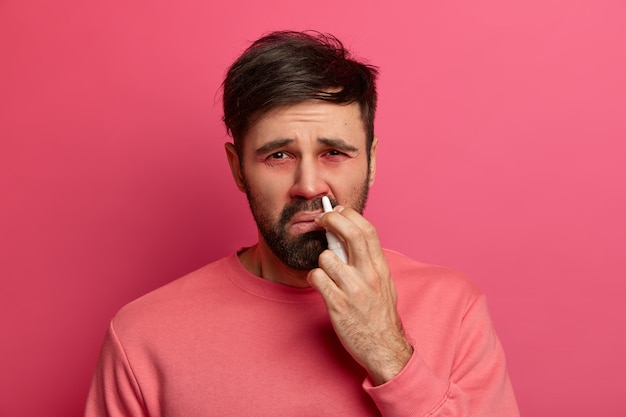 Un uomo malato sconvolto spruzza medicine per l'allergia al naso, ha preso raffreddore, soffre di rinite, ha occhi rossi e gonfi, vestito con abiti casual, posa contro il muro rosa. Concetto di trattamento della malattia.