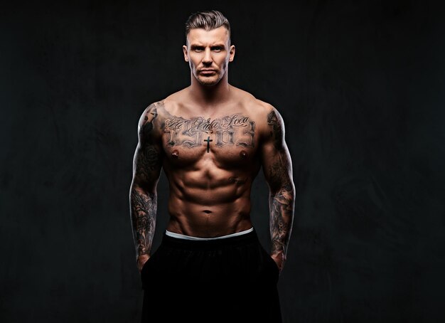 Un uomo muscoloso tatuato senza camicia con capelli alla moda in posa davanti alla telecamera su uno sfondo scuro.