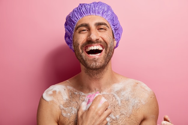 Un uomo nudo felicissimo si lava con il gel, ha il sapone sul corpo, tiene la spugna da bagno, indossa un berretto da doccia sulla testa, gode della freschezza, fa la doccia, sorride ampiamente, mostra i denti bianchi